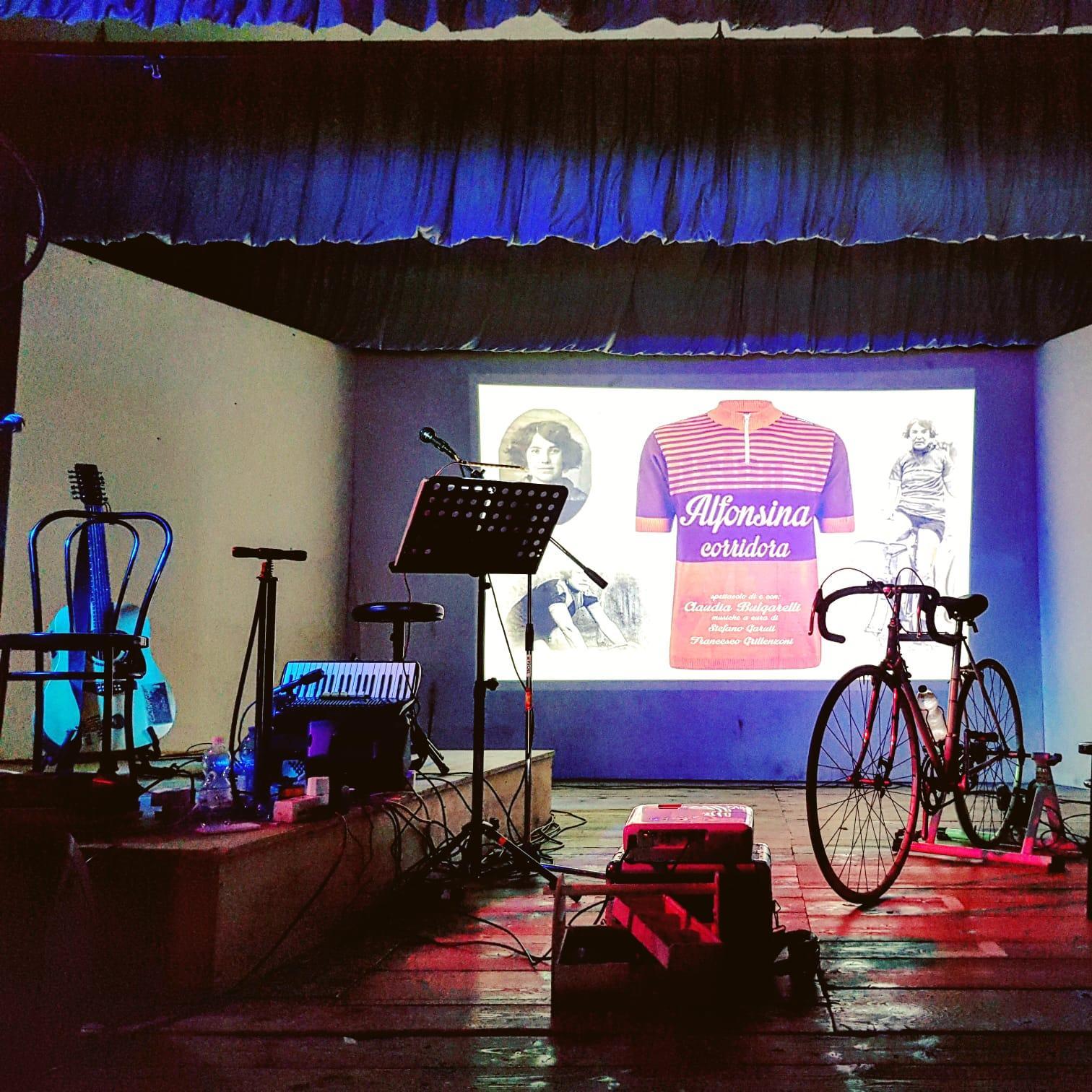 Palco allestito per Alfonsina, corridora con bicicletta, strumenti e titolo dello spettacolo proiettato.