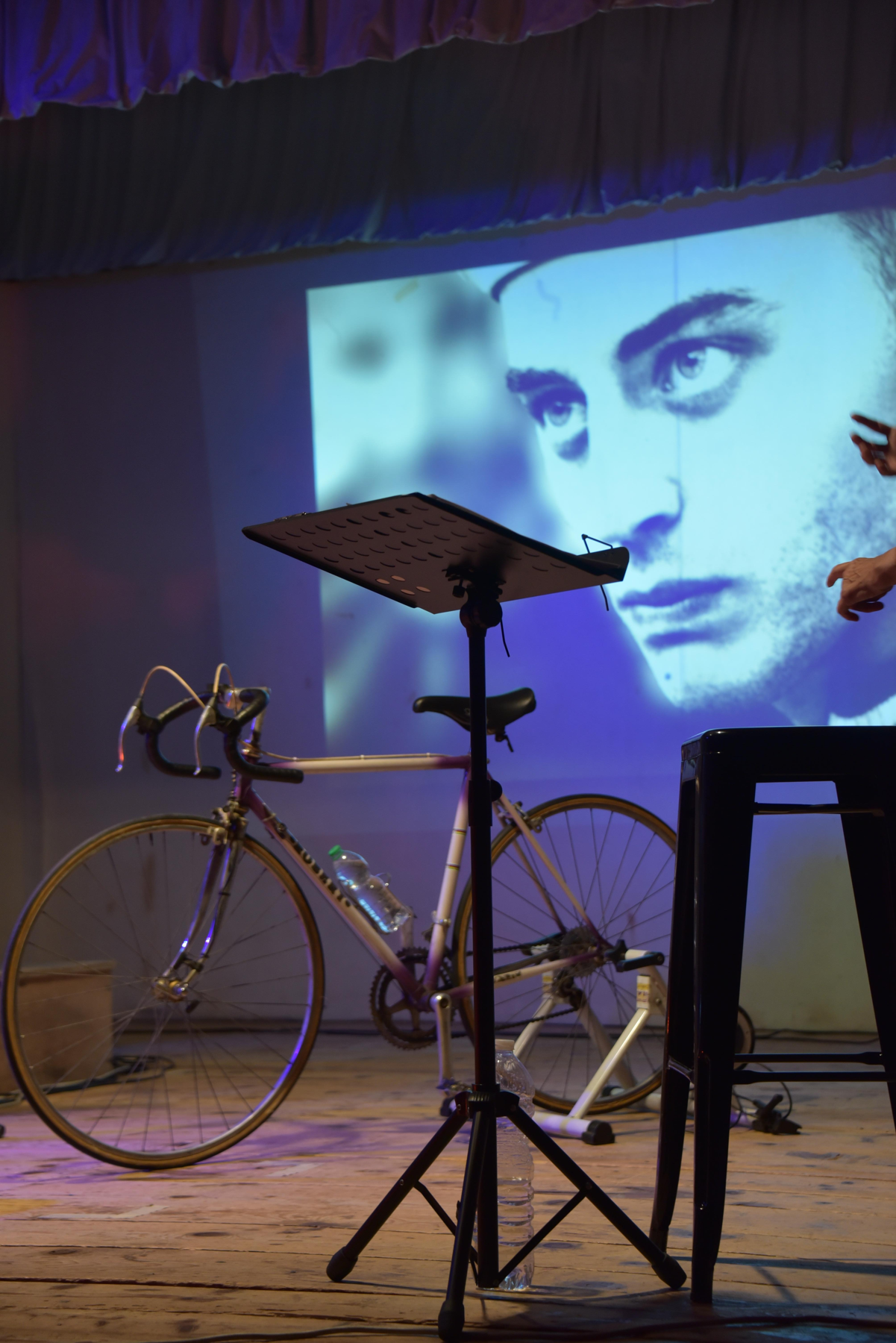 Bicicletta e leggio sul palco, davanti alla proiezione di un ritratto d’epoca.